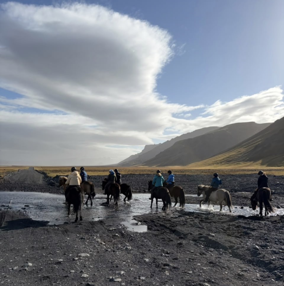 A line of riders on Icelandic horses crossing a shallow river under a wide cloudy sky