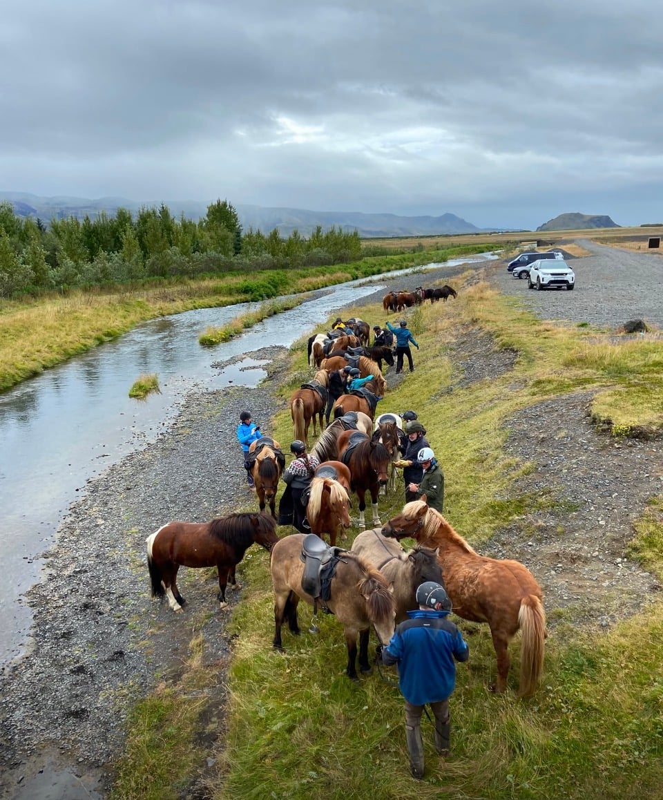 A line of Icelandic horses and riders gathered along a river during a multi-day riding tour